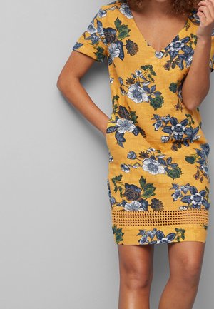 Femme portant une robe jaune à fleurs à manches courtes avec bordure en dentelle près de l'ourlet, une main dans la poche, sur un fond gris uni.