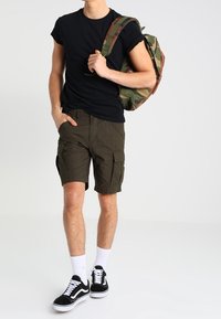 Černé tričko, olivově zelené cargo šortky s kapsami, bílé ponožky a černé skate boty; nese kamuflážový batoh.