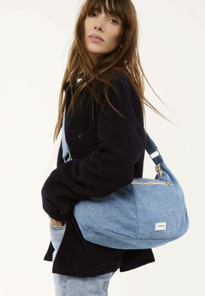 Helles blaues Denim-Umhängetasche mit geschwungener Form, goldener Reißverschluss und Logo-Patch. Die Tasche verfügt über einen breiten Tragegurt für zusätzlichen Komfort.