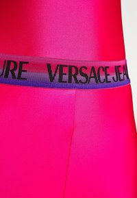 Felroze bodysuit met een contrasterende multikleurige tailleband met "VERSACE JEANS" in vetgedrukt zwart. Glad, glanzend textuur.