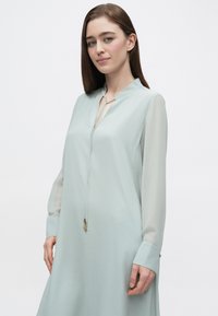 Hellminzgrünes Kleid mit Kragen, langen transparenten Ärmeln und einer zentralen Bindedetail. Es zeichnet sich durch eine glatte Textur und ein minimalistisches Design aus.