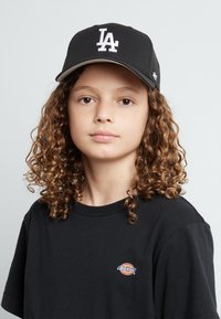 Cappellino da baseball nero con logo "LA" bianco, abbinato a una maglietta nera con un piccolo logo Dickies multicolore. Capelli ricci visibili.