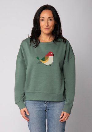 Femme aux cheveux foncés portant un sweat-shirt vert avec un motif abstrait d'oiseau et un jean bleu clair, se tenant devant un fond gris uni.