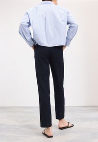 Chemise oversize à rayures bleues et blanches, vue de dos. Associée à un pantalon marine et des tongs noires. Tissu doux, coupe décontractée.