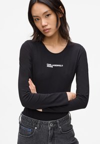 Černý dlouhý rukáv top s kulatým výstřihem a bílým logem "KARL LAGERFELD JEANS", nošený s tmavými džínami. Hladká textura.