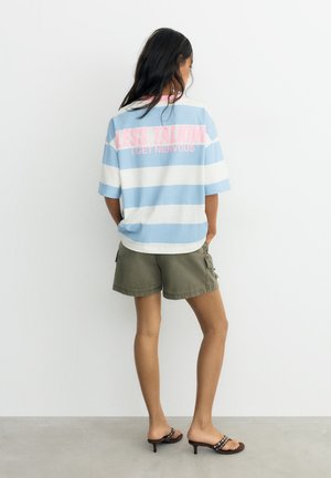 Junge Frau steht mit dem Rücken zur Kamera, trägt ein blau-weiß gestreiftes Shirt mit pinkem Text und olivgrüne Shorts mit Sandalen mit Absatz.