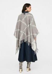 Poncho grigio con stampa del marchio ripetuta, frange lungo i bordi, indossato sopra una lunga gonna di denim e stivali alti fino al ginocchio con motivo.