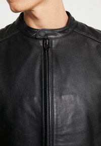 Giacca in pelle nera con colletto alto, zip frontale e una texture liscia con una sottile venatura.
