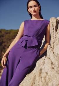 Lila ujjatlan ruha, testhez simuló felső résszel és nagy masnival a deréknál, sima textúrájú, lágy sziluettel, sziklás területen.