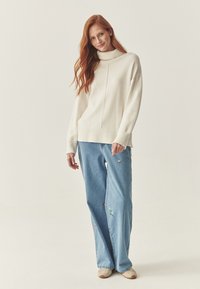 Crème coltrui met geribde manchetten en zoom, gecombineerd met lichtblauwe wijde jeans met kleurrijke bloemenborduursels.