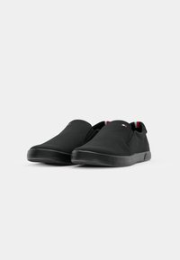 Paire de chaussures décontractées noires sans lacets avec semelles en caoutchouc et petits accents en tissu rouge, blanc et bleu sur la zone de la languette.