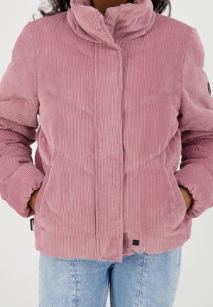 Vrouw draagt een roze corduroy gewatteerde jas met handen in de zakken en lichtblauwe spijkerbroek tegen een effen achtergrond.