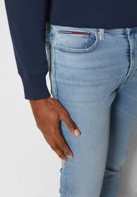 Prawa ręka spoczywa na kieszeni jasnoniebieskich dopasowanych dżinsów, ubrana w ciemnogranatowy top z długim rękawem na jednolitym jasnym tle.