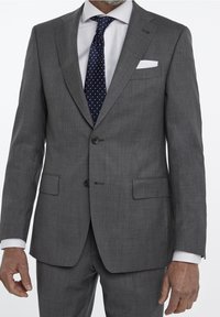 Costume grison avec fermeture à deux boutons, revers en pointe et poches latérales. Porté avec une chemise blanche et une cravate à pois bleu marine.
