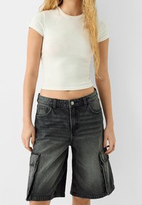Femme portant un crop top blanc à manches courtes et un short cargo en denim noir taille haute, debout devant un fond uni.