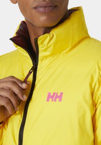 Žlutá bunda s vysokým límcem, na které je na prsou růžové logo, zapínání na zip a kontrastní vínové podšívání viditelné na límci.