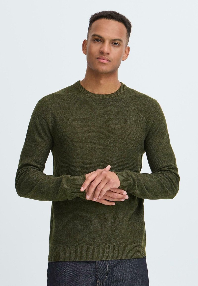 Olijfgroene trui van textiel met een structuur, met een ronde halslijn en geribbelde boorden. Model staat met gekruiste armen en draagt een donkere spijkerbroek.