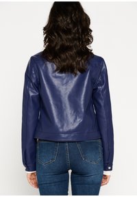 Giacca in finta pelle blu con design aderente, chiusura con zip e polsini a costine, abbinata a jeans blu scuro con tasche posteriori.