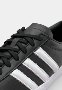 Melnas ādas sneakers ar baltu trīsu strīpu dizainu, apaļu purngali, plakanu melnu šņoru un strukturētu izšuvumu uz virsmas.