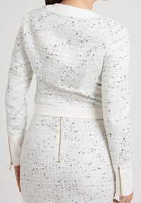 Vit texturerad jacka med svarta fläckar, med zippered nederkant och ärmslut. Slät krage, figursydd design och distinkt tygmönster.