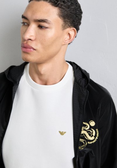 Casaco leve preto com fecho éclair e bordado dourado, sobreposto a uma t-shirt branca de gola redonda com um pequeno logótipo dourado.