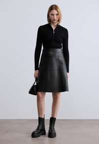 Μαύρο ριμπ πλεκτό τοπ με φερμουάρ σε συνδυασμό με μαύρη δερμάτινη φούστα σε σχήμα Α. Το μοντέλο φοράει μαύρα χοντρά μποτάκια μέχρι τον αστράγαλο και κρατά μια μικρή τσάντα χειρός.