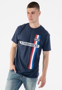 Navy blue cotton t-shirt featuring a white "Knebworth" logo and a vertical red, white, and blue stripe on the left side. Short sleeves, round neck.