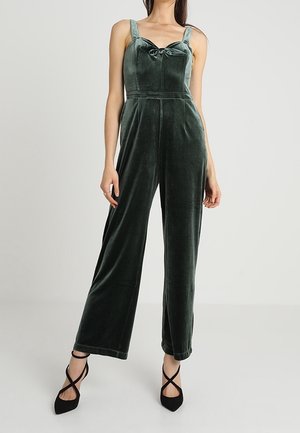 Velvet green jumpsuit with wide legs, a fitted bodice, and bow detail at the neckline. Features adjustable straps and a soft texture.