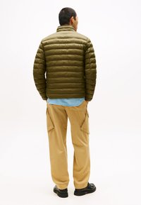 Grøn pufferjakke med vandret quiltet mønster, høj krave og letvægtsdesign. Kombineret med beige cargo bukser og sorte sko.