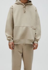Sweat à capuche beige avec une poche avant, une capuche à cordon ajustable, une texture douce et une étiquette discrète sur le côté, associé à un pantalon beige assorti.