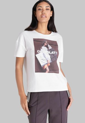 Vrouw met lang donker haar draagt een wit T-shirt met een grote foto van een model dat een zilveren clutch vasthoudt en de tekst "CHOCOLATE BITE."