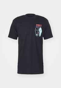Tricou negru din bumbac, cu un imprimeu grafic al picioarelor și textul "OBEY" în accente de roșu și albastru. Gât scund și mâneci scurte.