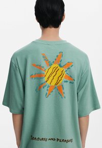 Green oversized t-shirt featuring a textured sun graphic in yellow and orange on the back, with "Treasures and Pleasures" printed below.