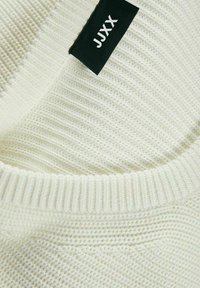 Sweater branco de malha com textura canelada e gola redonda. Apresenta um rótulo preto com o texto "JJXX" na lateral.