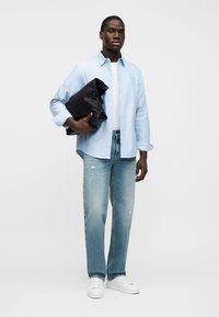 Άνδρας με ανοιχτόχρωμο γαλάζιο πουκάμισο και τζιν κρατά μια μεγάλη μαύρη τσάντα χειρός μπροστά σε λευκό φόντο.