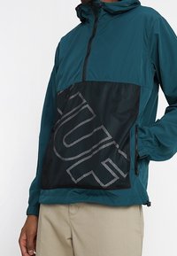 Tyrkysová lehká bunda s černou síťovinovou panelem, obsahující velké bílé písmo, nastavitelnou kapuci a boční kapsy, kontrastní textury látek.