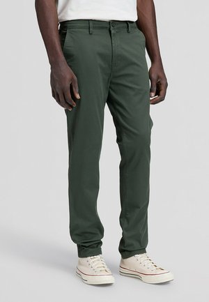 Mann trägt dunkelgrüne Hose mit geradem Bein und weiße Leinenschuhe und steht vor einem einfarbigen Hintergrund.