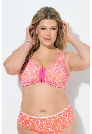 Kobieta plus size poprawiająca włosy, ubrana w różowy i pomarańczowy biustonosz w panterkę i pasującą bieliznę, stojąca na białym tle.