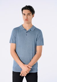 Camisa polo de manga corta azul con tela texturizada, con tapeta de dos botones y cuello. Lleva pantalones negros y está de pie contra un fondo claro.