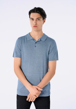 Blå kortærmet polo-shirt med tekstureret stof, to-knaps placket og krave. Båret med sorte bukser, stående mod en lys baggrund.