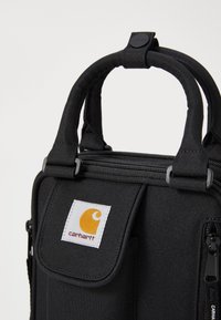 Sac en tissu noir avec doubles poignées, fermetures éclair et poche avant à rabat. Présente une étiquette blanche avec un logo orange. Texture textile solide.