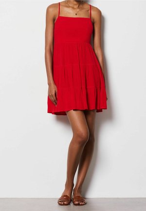 Kobieta ubrana w czerwoną sukienkę na ramiączkach z falbanami, z cienkimi szelkami, złoty naszyjnik i brązowe płaskie sandały, stoi przy białej ścianie.