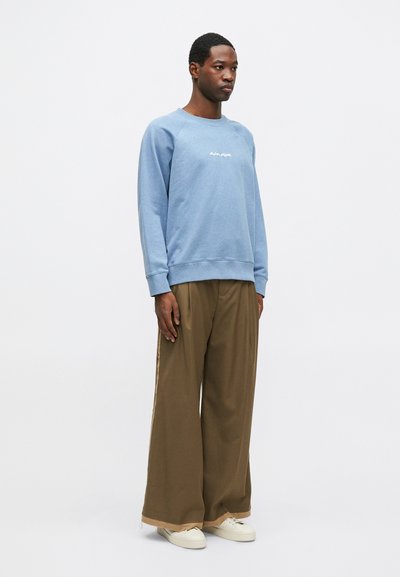 Sudadera azul claro con texto blanco, combinada con pantalones de pierna ancha marrones y zapatillas blancas. Tela suave, corte relajado y puños acanalados.