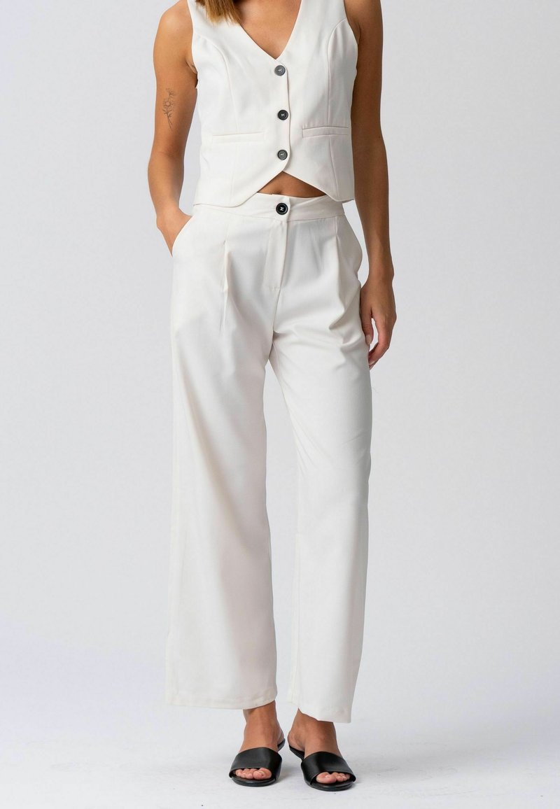 Femme portant un gilet blanc sans manches à boutons, un pantalon blanc à jambes larges et des sandales noires ouvertes à l'avant, debout avec une main dans la poche.