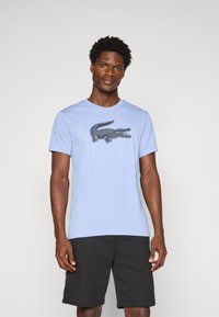 Homem a usar uma t-shirt azul-clara com um grande logótipo de crocodilo escuro e calções pretos, de pé contra um fundo claro.