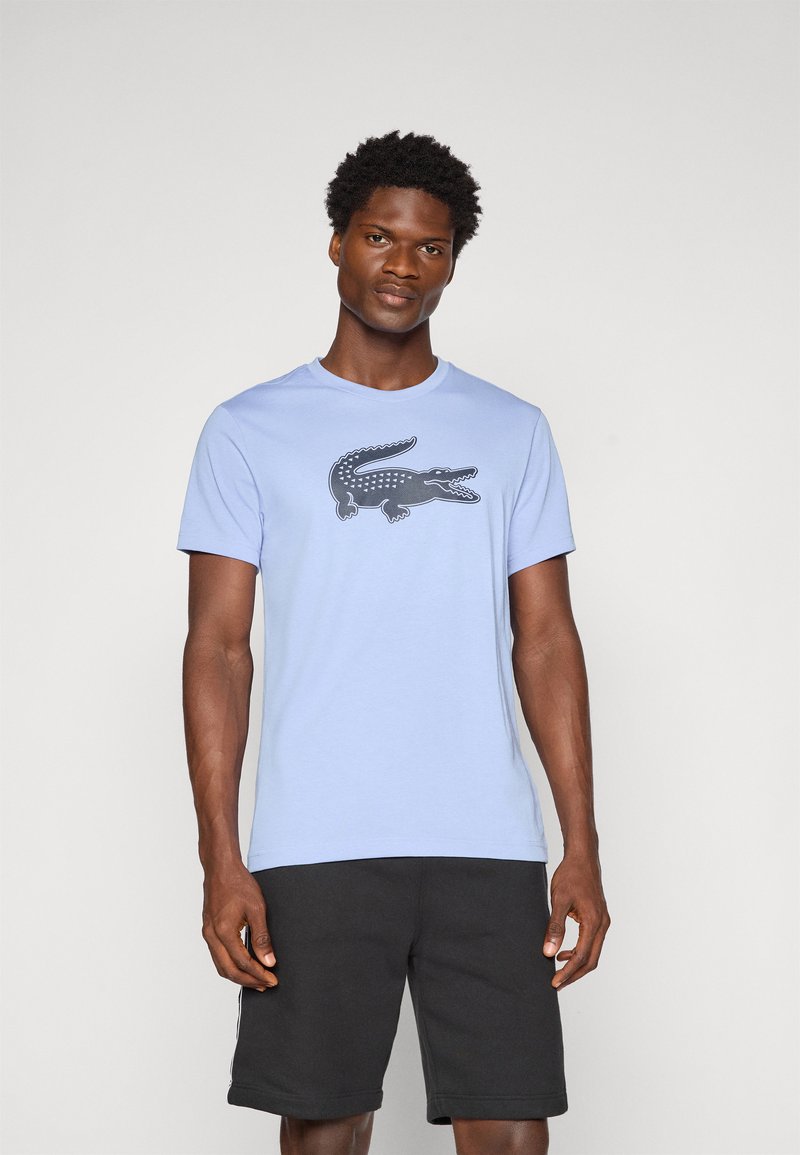 Homem a usar uma t-shirt azul-clara com um grande logótipo de crocodilo escuro e calções pretos, de pé contra um fundo claro.