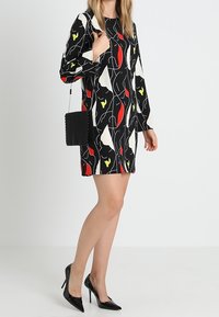 Mustapuku, jossa on abstrakteja punaisia, valkoisia ja keltaisia kuvioita, pitkät hihat, yhdistettynä pieneen mustaan helmibagniin ja mustiin korkokenkiin.