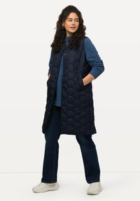 Marinblå pufferväst med hexagonal quiltad design, två sidofickor, tryckknappar, bärs över en blå tröja och matchas med mörka jeans och vita sneakers.