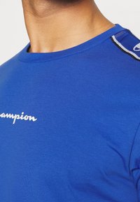 Modrá bavlněná tričko s kulatým výstřihem, s bílým logem "Champion" a černými akcenty na rameni. Měkká textura.