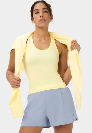 Vrouw met gele tanktop en lichtblauwe sportieve korte broek houdt een bijpassende gele trui over haar schouders.